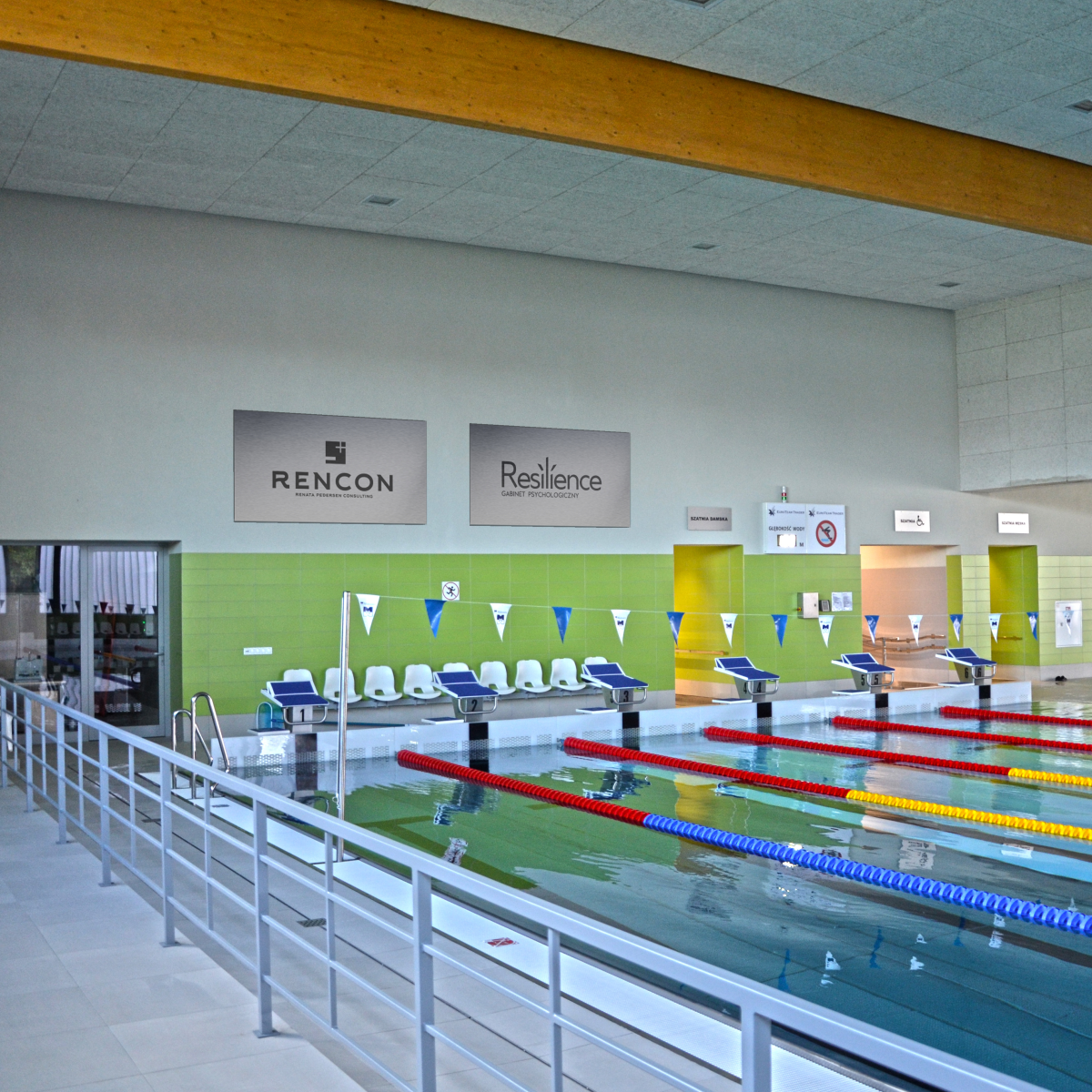 Płyta basenowa z widokiem na sześć torów pływackich. Na ścianie umieszczone są przykładowe reklamy do wynajęcia.